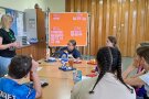 Kinder sitzen an Tisch, Frau steht daneben