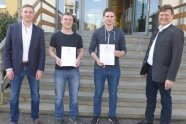 4 Personen stehen nebeneinander vor Treppe eines Gebäudes, zwei halten Urkunden